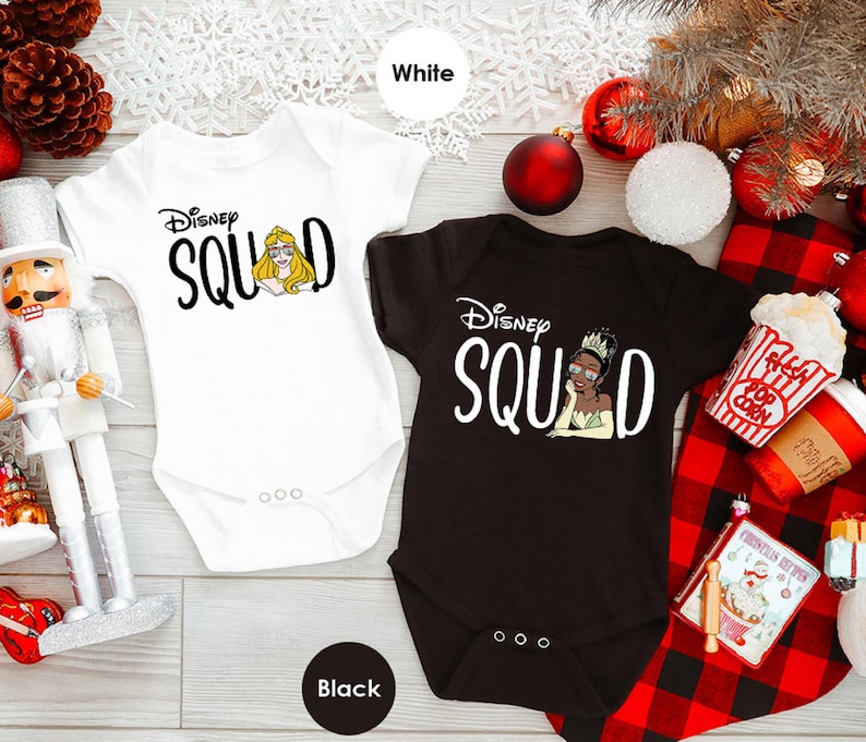 May include: Two baby onesies, one white and one black, with the text "Disney Squad". The white onesie features a graphic of Princess Aurora, and the black onesie features Princess Tiana. Both have short sleeves and three snaps at the bottom.
