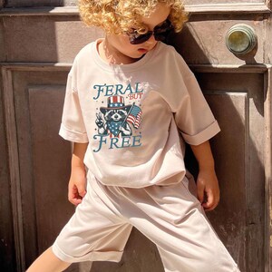May include: A child wearing a light pink t-shirt and shorts set. The shirt features a raccoon graphic wearing a hat and holding an American flag, with the words "FERAL BUT FREE". The child is also wearing sunglasses.