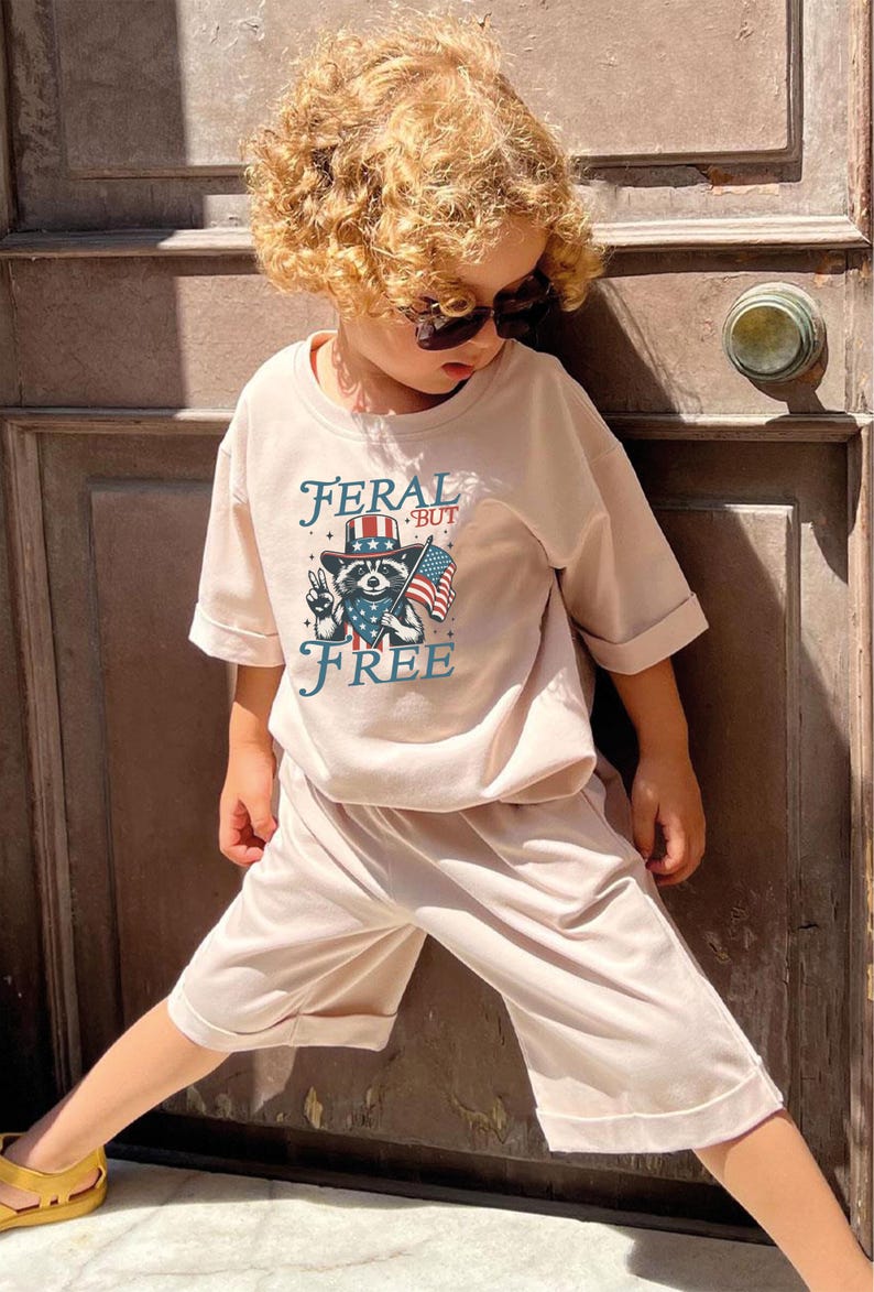 May include: A child wearing a light pink t-shirt and shorts set. The shirt features a raccoon graphic wearing a hat and holding an American flag, with the words "FERAL BUT FREE". The child is also wearing sunglasses.