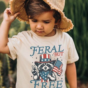 May include: A tan t-shirt with the words "FERAL BUT FREE" in blue script. The shirt features a raccoon wearing a patriotic hat and bandana, holding an American flag and making a peace sign. The child is wearing a straw hat.