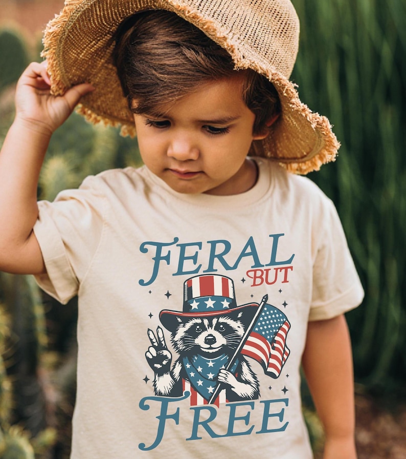 May include: A tan t-shirt with the words "FERAL BUT FREE" in blue script. The shirt features a raccoon wearing a patriotic hat and bandana, holding an American flag and making a peace sign. The child is wearing a straw hat.
