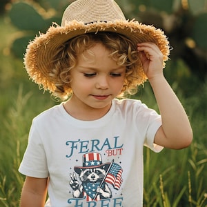 May include: A young child wearing a straw hat and a white t-shirt with the words "FERAL BUT FREE" and a raccoon graphic holding an American flag. The raccoon is wearing a patriotic hat and bandana.