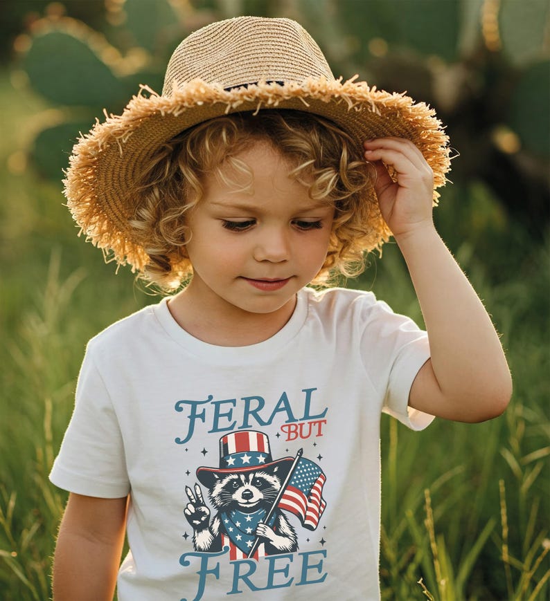 May include: A young child wearing a straw hat and a white t-shirt with the words "FERAL BUT FREE" and a raccoon graphic holding an American flag. The raccoon is wearing a patriotic hat and bandana.