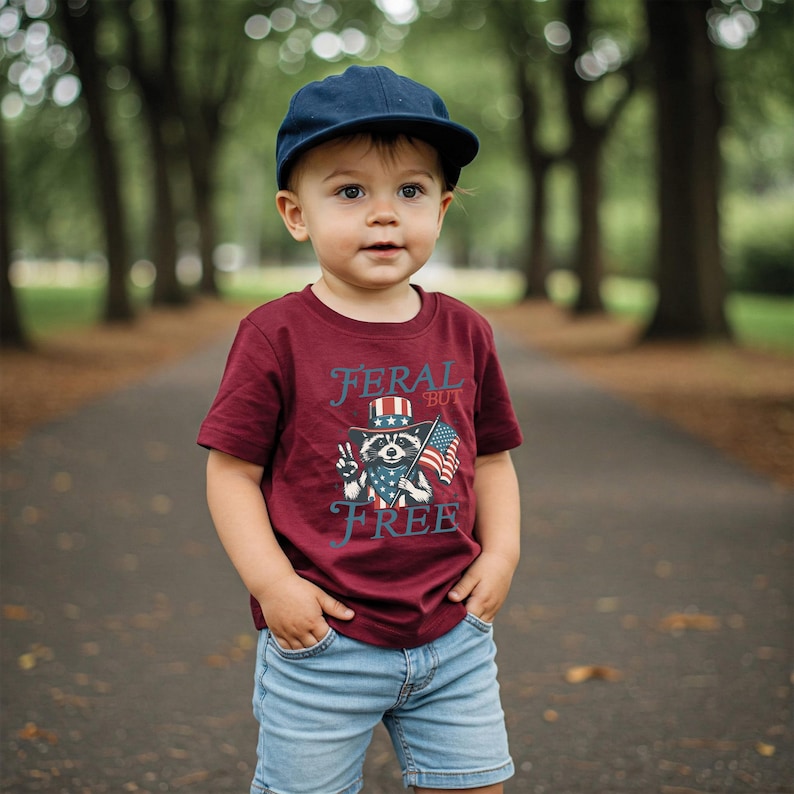 May include: A young child wearing a maroon t-shirt, blue jeans, and a navy blue cap. The t-shirt features a raccoon graphic with an American flag and the text "FERAL BUT FREE". The child is standing on a paved path with trees in the background.