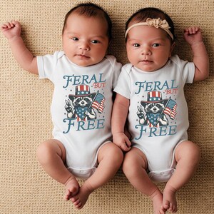 May include: Two babies wearing white onesies with a raccoon graphic, an American flag, and the text "FERAL BUT FREE". One baby has a beige bow headband. The babies are lying on a beige surface.