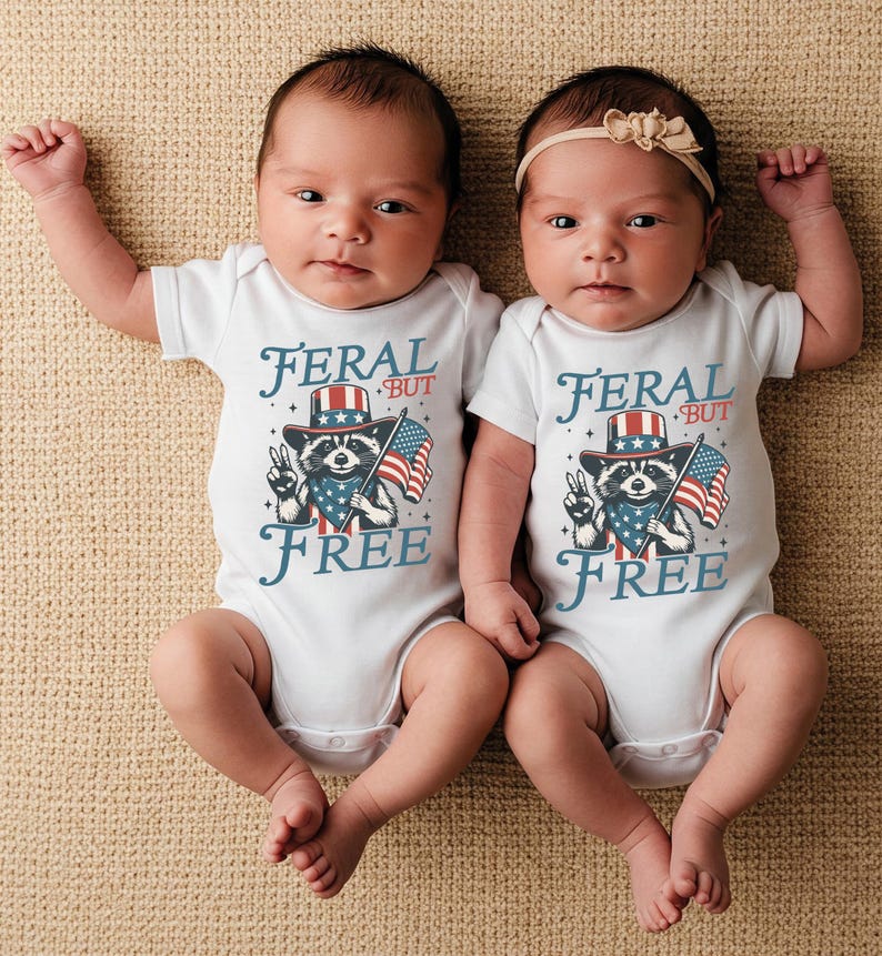 May include: Two babies wearing white onesies with a raccoon graphic, an American flag, and the text "FERAL BUT FREE". One baby has a beige bow headband. The babies are lying on a beige surface.