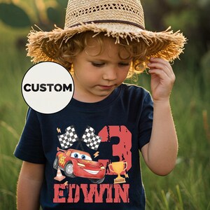 Puede incluir: Un ni&ntilde;o con una camiseta azul marino con un gr&aacute;fico de coche de carreras rojo, el n&uacute;mero 3, un trofeo y el nombre "EDWIN". La camiseta tambi&eacute;n presenta banderas a cuadros y un sombrero de paja.