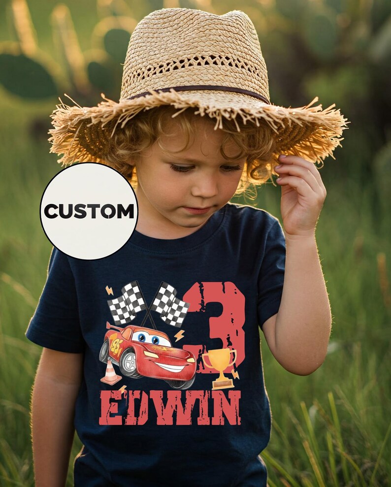 Puede incluir: Un ni&ntilde;o con una camiseta azul marino con un gr&aacute;fico de coche de carreras rojo, el n&uacute;mero 3, un trofeo y el nombre "EDWIN". La camiseta tambi&eacute;n presenta banderas a cuadros y un sombrero de paja.