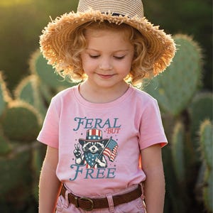 May include: A young child wearing a pink t-shirt with the words "FERAL BUT FREE" and a graphic of a raccoon in a patriotic hat holding an American flag. The child is also wearing a straw hat and brown belt.