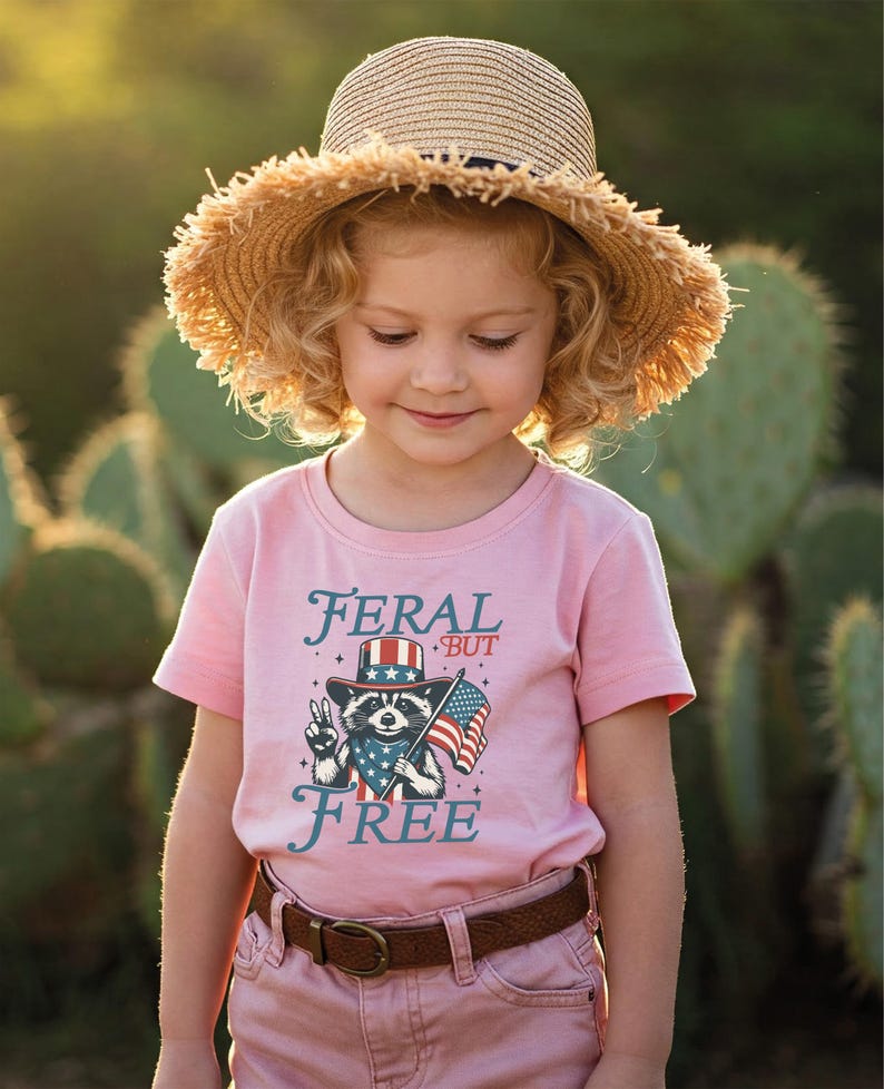 May include: A young child wearing a pink t-shirt with the words "FERAL BUT FREE" and a graphic of a raccoon in a patriotic hat holding an American flag. The child is also wearing a straw hat and brown belt.