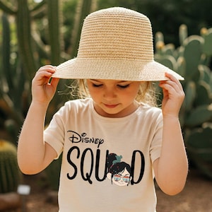 May include: A young person wearing a beige t-shirt with the words "Disney Squad" in black script and a graphic of a character wearing sunglasses. They are also wearing a wide-brimmed straw hat.