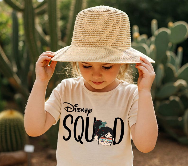 May include: A young person wearing a beige t-shirt with the words "Disney Squad" in black script and a graphic of a character wearing sunglasses. They are also wearing a wide-brimmed straw hat.