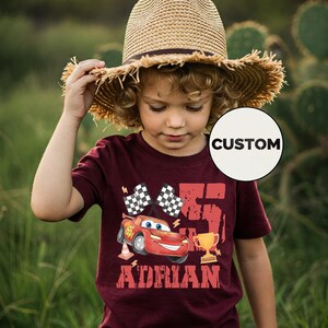 Puede incluir: Una camiseta granate con un coche de carreras de dibujos animados, banderas a cuadros, un trofeo y el nombre "Adrian". El n&uacute;mero "75" est&aacute; impreso en grande. Un ni&ntilde;o lleva la camiseta y un sombrero de paja.