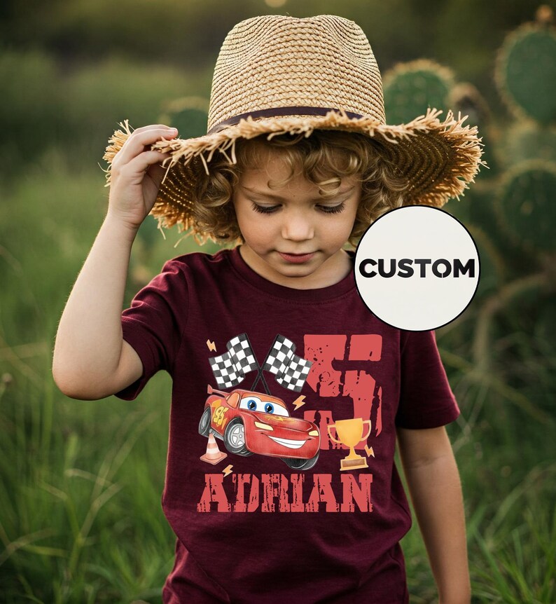Puede incluir: Una camiseta granate con un coche de carreras de dibujos animados, banderas a cuadros, un trofeo y el nombre "Adrian". El n&uacute;mero "75" est&aacute; impreso en grande. Un ni&ntilde;o lleva la camiseta y un sombrero de paja.