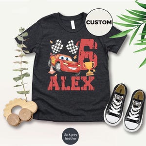 Puede incluir: Camiseta gris oscuro jaspeado con un gr&aacute;fico de coche de carreras rojo, el n&uacute;mero 6, un trofeo, banderas a cuadros y el nombre "ALEX" en letras rojas. Zapatillas deportivas negras y blancas colocadas junto a la camiseta.