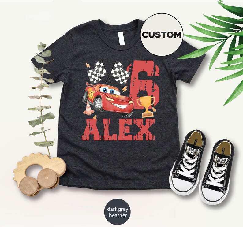 Puede incluir: Camiseta gris oscuro jaspeado con un gr&aacute;fico de coche de carreras rojo, el n&uacute;mero 6, un trofeo, banderas a cuadros y el nombre "ALEX" en letras rojas. Zapatillas deportivas negras y blancas colocadas junto a la camiseta.