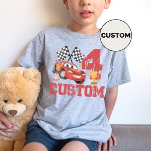 Puede incluir: Una camiseta gris con un coche de carreras de dibujos animados rojo, banderas a cuadros, el n&uacute;mero 4 y un trofeo. La palabra "CUSTOM" est&aacute; impresa en rojo debajo del coche. Un c&iacute;rculo blanco con la palabra "CUSTOM" est&aacute; en la camiseta.