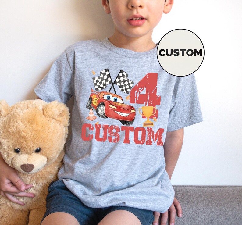 Puede incluir: Una camiseta gris con un coche de carreras de dibujos animados rojo, banderas a cuadros, el n&uacute;mero 4 y un trofeo. La palabra "CUSTOM" est&aacute; impresa en rojo debajo del coche. Un c&iacute;rculo blanco con la palabra "CUSTOM" est&aacute; en la camiseta.