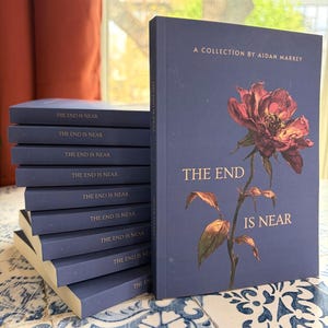 May include: A stack of books with navy blue covers. The book on the right has the title "THE END IS NEAR" with a floral illustration. The author's name, Aidan Markey, is also visible. The books are on a blue and white patterned surface.