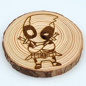 May include: A wooden coaster with a laser-engraved image of a cartoon Deadpool character. The character is wearing a red and black suit and has a mask on. The coaster is round and has a natural wood finish.