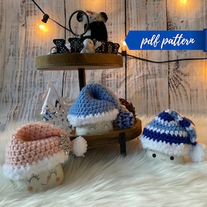 May include: Three crocheted snowman hats in pink, blue, and blue and white stripes. The hats are on top of white mugs with smiling faces. The mugs are on a tiered wooden tray with a wooden background.