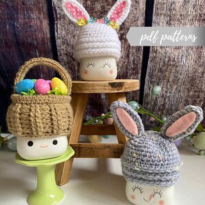 May include: Three white ceramic mugs with bunny ears and crocheted hats. The mugs have a smiling face and are decorated with pastel colored Easter eggs. The hats are white, gray, and brown. The text "pdf patterns" is visible in the image.