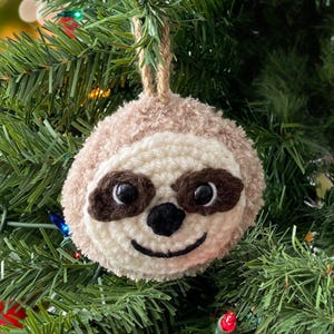 May include: A crocheted sloth ornament with brown eyes, a black nose, and a brown mouth. The ornament is hanging on a green Christmas tree.