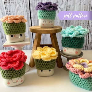 May include: Six crocheted flower mug toppers in various colors, including purple, peach, teal, yellow, pink, and red. Each topper features a green crocheted base and a smiling face.