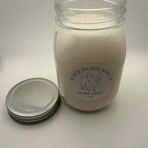 May include: A clear glass jar filled with white lard, with a silver lid next to it. The jar has a label that reads "Black Horse Ranch Pork Lard 1 LB" with a pig illustration. The jar is on a white surface.