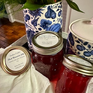 May include: Two glass jars of Southern Dabblers Black Muscadine Jelly. The jars are on a white cloth with a blue and white patterned vase in the background. The labels on the jars read "Southern Dabblers Black Muscadine Jelly".
