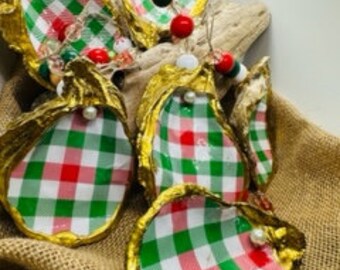 May include: Handmade oyster shell Christmas ornaments. Each shell is painted gold around the edges and features a red, green, and white plaid pattern. The ornaments are adorned with beads and pearls, and are displayed on a piece of driftwood.