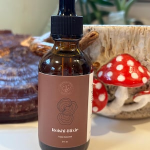 以下が含まれることがあります： ドロップトップと「Reishi Elixir Triple Extracted 2 fl. oz.」と書かれたラベルが付いた茶色のガラス瓶。瓶は、キノコとセラミック製のマグカップを背景に、白い表面の上に置かれています。
