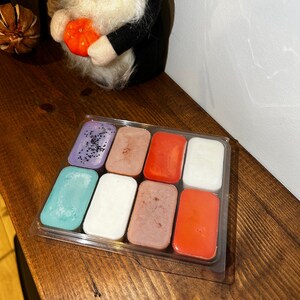 以下が含まれることがあります： さまざまな色の長方形のワックスメルトが8つ入った透明なプラスチック容器。メルトには、紫、ティール、白、茶色、オレンジの色合いが含まれます。一部のメルトには装飾的なグリッターが付いています。木製の表面に置かれています。