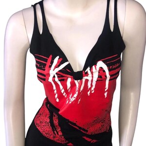 May include: Black tank top with a deep V-neck and double strap detail. The top features a red and black graphic design with the word "Korn" in white, a band logo.