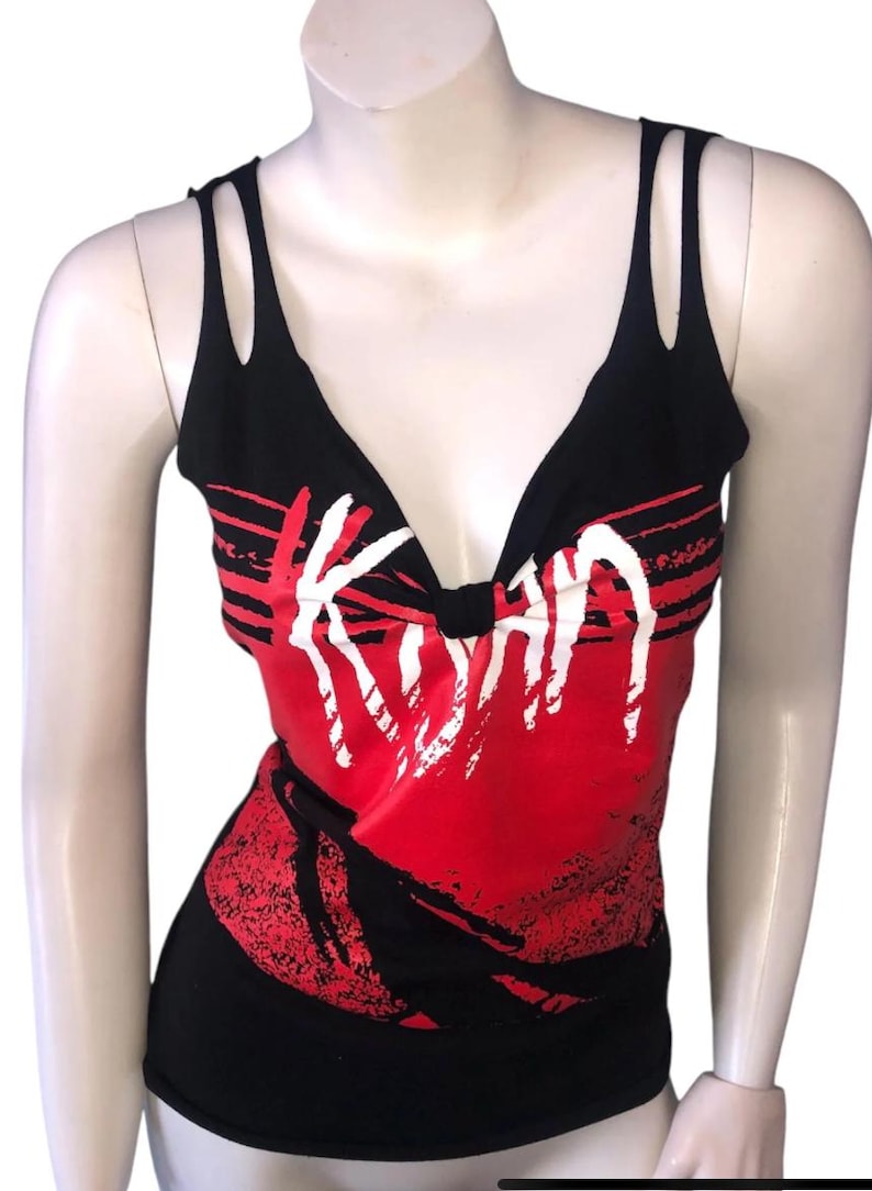 May include: Black tank top with a deep V-neck and double strap detail. The top features a red and black graphic design with the word "Korn" in white, a band logo.