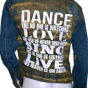 Op de afbeelding: Een blauwe denimjas met gouden verfspatten en witte tekst op de rug die luidt "Dance like no one is watching, Love like you've never been hurt, Sing like no one is listening, Live like heaven is on earth."