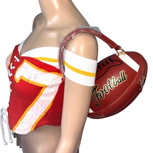 May include: Red and white football-themed corset top with the number 7 in yellow. A matching red and white football-shaped handbag with the word "Football" in gold script is held by a mannequin.