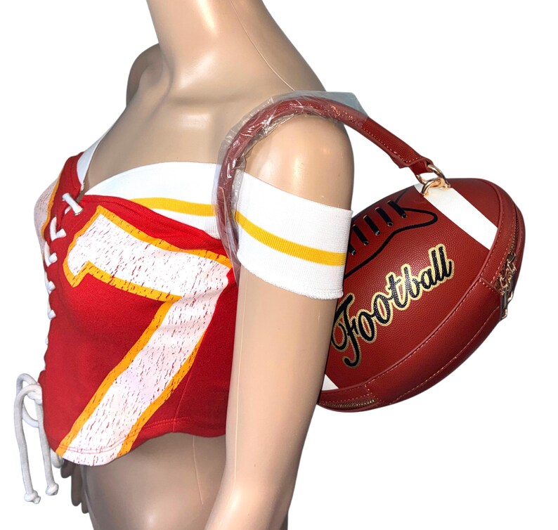 May include: Red and white football-themed corset top with the number 7 in yellow. A matching red and white football-shaped handbag with the word "Football" in gold script is held by a mannequin.