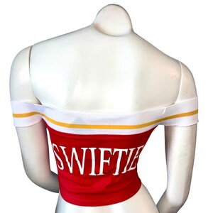 May include: Red off-the-shoulder crop top with the word "SWIFTIE" in white block letters. The top has a white band with a yellow stripe along the top and around the sleeves. The top is displayed on a white mannequin.