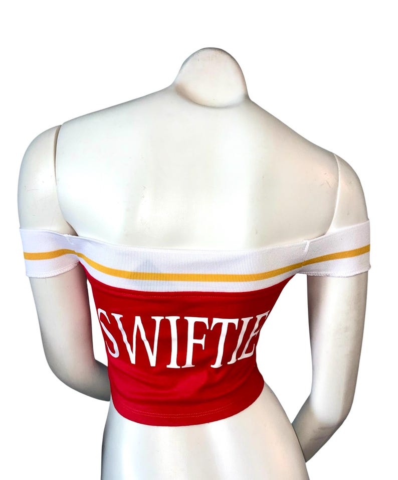 May include: Red off-the-shoulder crop top with the word "SWIFTIE" in white block letters. The top has a white band with a yellow stripe along the top and around the sleeves. The top is displayed on a white mannequin.