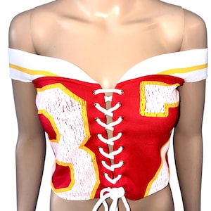 May include: Red and white off-the-shoulder corset top with the number 83 in white and yellow. The top has a lace-up front and white and yellow trim along the neckline.