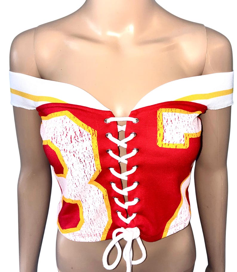 May include: Red and white off-the-shoulder corset top with the number 83 in white and yellow. The top has a lace-up front and white and yellow trim along the neckline.