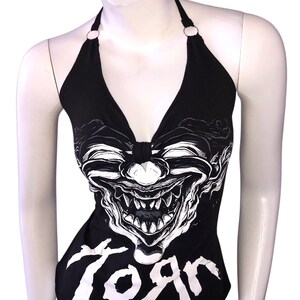 K&ouml;nnte beinhalten: Schwarzes Halstuch-Top mit einem wei&szlig;en Grafikdruck eines Clownsgesichts und dem Wort "Korn" auf der Vorderseite.