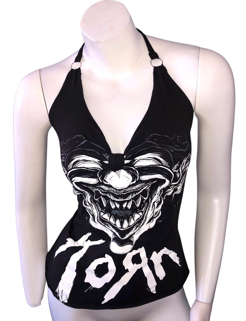 K&ouml;nnte beinhalten: Schwarzes Halstuch-Top mit einem wei&szlig;en Grafikdruck eines Clownsgesichts und dem Wort "Korn" auf der Vorderseite.
