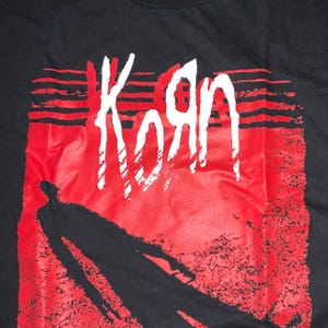 May include: Black t-shirt with the band name "Korn" in white, distinctive lettering set against a red backdrop. The design incorporates a silhouetted figure. The shirt has a crew neck and a tag at the top with the band's name and size details.