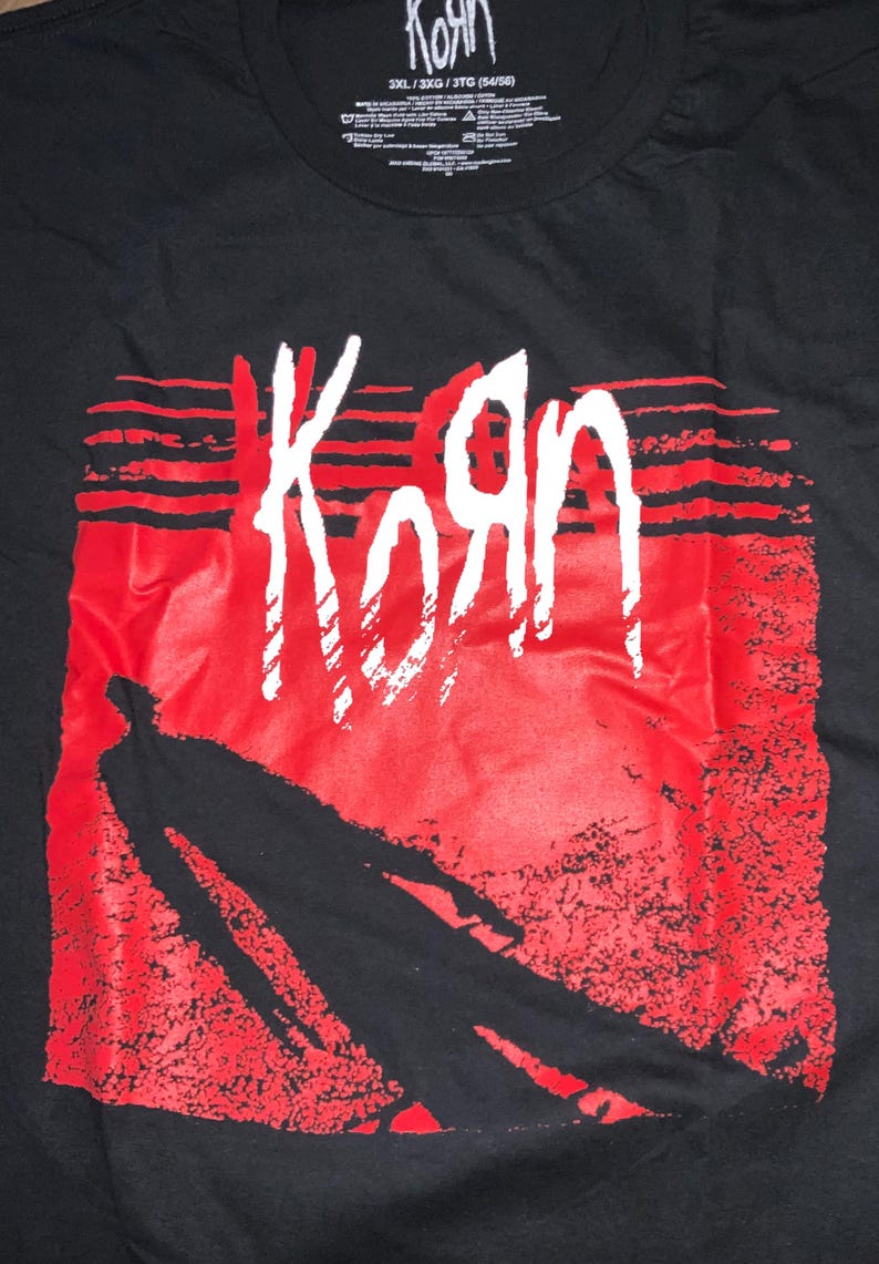May include: Black t-shirt with the band name "Korn" in white, distinctive lettering set against a red backdrop. The design incorporates a silhouetted figure. The shirt has a crew neck and a tag at the top with the band's name and size details.