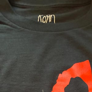 May include: Black t-shirt featuring the band name "Korn" in white script at the collar and large red lettering across the front. The shirt is made of a dark, soft fabric.
