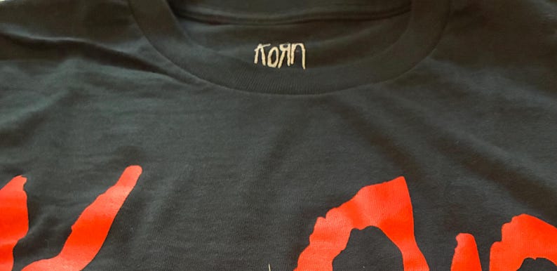 May include: Black t-shirt featuring the band name "Korn" in white script at the collar and large red lettering across the front. The shirt is made of a dark, soft fabric.