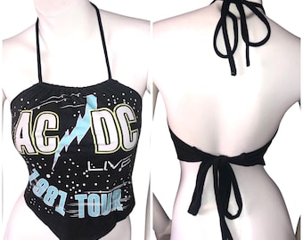 May include: Black halter top featuring the AC/DC Live 1981 Tour graphic in white, yellow, and blue. The top ties at the neck and back. The front of the top has a triangular shape.
