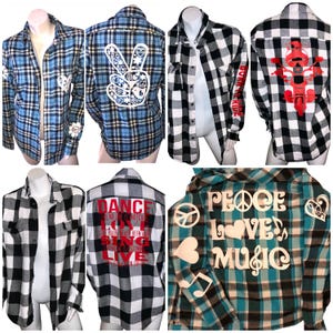 May include: Collection of plaid flannel shirts in various colors, including blue, black, and teal. Some shirts feature white graphics like peace signs, hearts, and musical notes. Others have text like "DANCE" and "PEACE LOVE MUSIC".